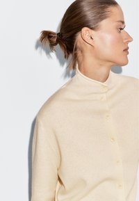 Beige stickad cardigan med hög krage och framknäppning. Mjuk textur och minimalistisk design, med sex små knappar.