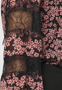 Haut transparent noir avec des motifs floraux roses, agrémenté de détails en dentelle noire sur les manches. Le tissu texturé crée un contraste dans le design.