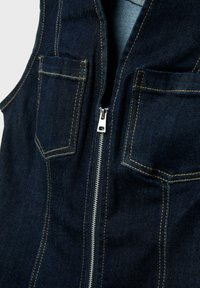 Tmavě modrá džínová vesta s předním stříbrným zapínáním na zip a dvěma kapsami na prsou s žlutým prošíváním.