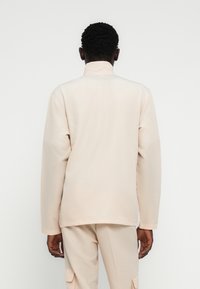 Ανοιχτό μπεζ μπουφάν με ψηλό γιακά, μακριά μανίκια και λεία υφή· απεικονίζεται από πίσω με ίσια κοπή και ελάχιστες λεπτομέρειες.