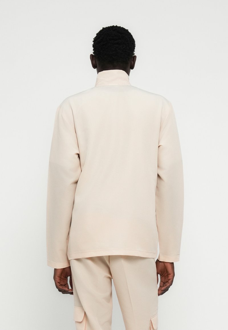 Veste beige clair avec un col montant, des manches longues et une texture lisse ; présentée de dos avec une coupe droite et des détails minimaux.