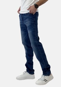 Jeans de denim azul oscuro con un corte ajustado, que presentan un ligero desgaste y arrugas. Combinados con zapatillas blancas y una camiseta blanca básica.