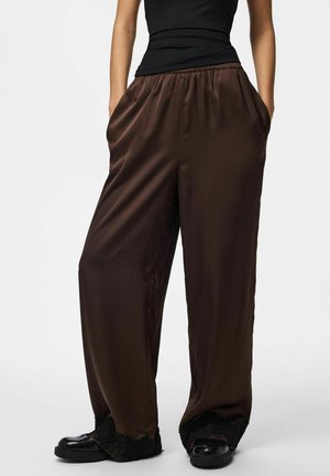 Osoba nosząca luźne, błyszczące ciemnobrązowe spodnie o szerokich nogawkach z koronkową lamówką na dole, czarne buty oraz dopasowany czarny bezrękawnik.
