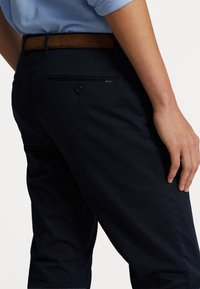 Pantaloni di cotone blu navy con una texture liscia, dotati di una tasca posteriore con chiusura a bottone e una cintura in pelle marrone.