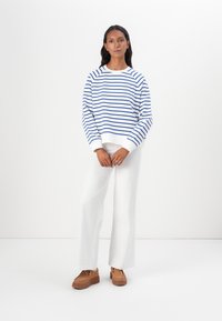 Felpa bianca con righe orizzontali blu, scollatura rotonda e polsini a costo. Abbinata a pantaloni bianchi a gamba larga e scarpe con zeppa color sabbia.