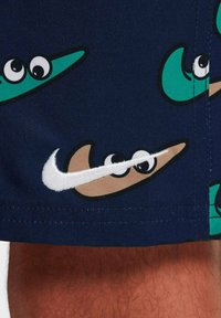 Bañadores de natación en color azul marino con patrones de peces de dibujos animados en verde y beige, contorneados en negro; cuenta con un destacado logotipo de Nike en blanco bordado en un lateral.