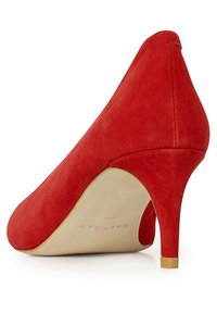 Punainen mokkanahkainen korkokenkä, jossa on terävä kärki ja kapea stilettikorko. Kenkässä on sileä pinta ja beiget pohjat, joissa on hienovarainen brändäys.