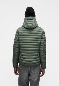 Vihreä toppatakki hupulla, jossa tikattu muotoilu, sileä pinta ja joustavat hihansuut, harmaiden farkkujen kanssa takaa päin nähtynä.