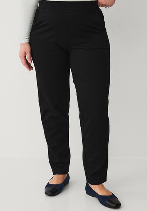 Person iført sorte, slim-fit bukser og marineblå ballet flats med sorte tåkapper, stående mod en ensfarvet baggrund.