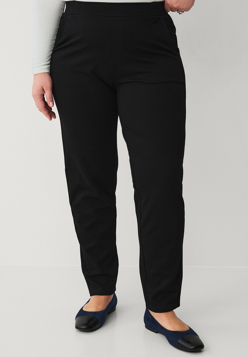 Person iført sorte, slim-fit bukser og marineblå ballet flats med sorte tåkapper, stående mod en ensfarvet baggrund.