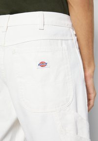 Pantalon blanc en tissu texturé, avec une poche au dos portant un patch logo "Dickies" coloré et des coutures plates.