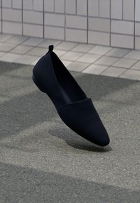 Zwarte platte schoen met een gladde textuur, spitse neus en minimalistisch ontwerp. Bevat een kleine hak en een lus aan de achterkant voor eenvoudig aan- en uittrekken.