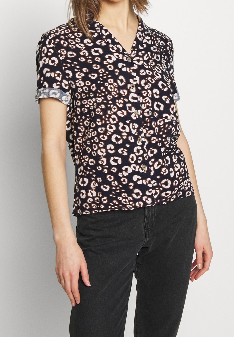 Personne portant une chemise noire à manches courtes boutonnée avec un motif léopard blanc et brun et un pantalon noir, debout devant un fond uni.
