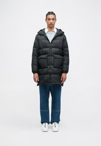 Giacca a vento trapuntata nera con cappuccio, dotata di zip frontale, due tasche sul petto e orli regolabili, abbinata a jeans a gamba larga blu e sneakers bianche.