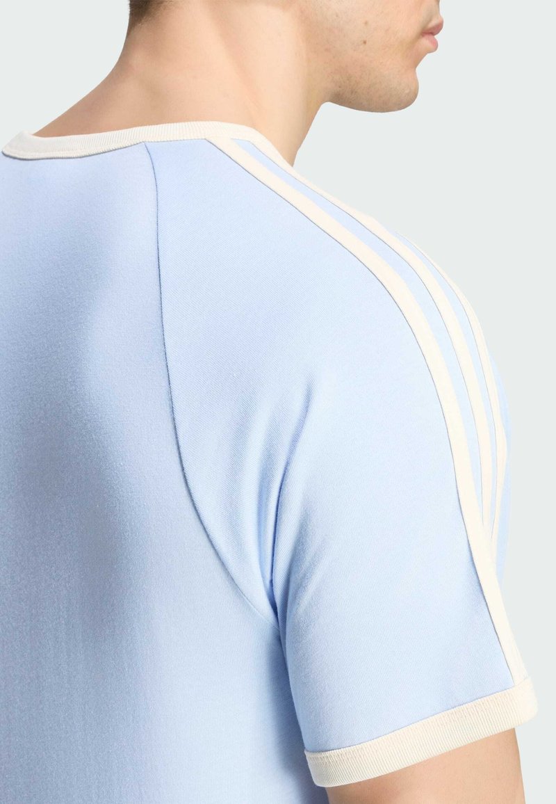 Uomo che indossa una camicia azzurro chiaro a maniche corte con rifiniture bianche e tre strisce bianche sulla spalla, mostrato di spalle e di lato.
