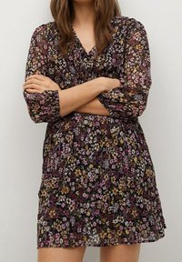 Květinové šaty v černé barvě, vyrobené z průhledné tkaniny s růžovými, fialovými, žlutými a bílými květy, s dlouhými rukávy a volným střihem.