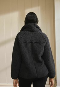 Veste en polaire gris foncé avec un col montant, une surface texturée et des épaules tombantes. Matière douce et confortable sans fermetures visibles ni motifs.