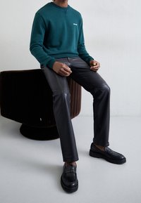 Niebieskozielona bluza z małym białym logo, zestawiona z czarnymi skórzanymi spodniami i czarnymi butami. Model siedzi na okrągłym, brązowym, teksturowanym fotelu.