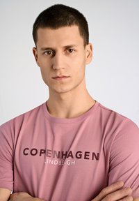 Rosa bomullströja med "COPENHAGEN LINDBERGH" tryckt i mörkbruna och vita bokstäver. Enkel rund halsringning och korta ärmar.