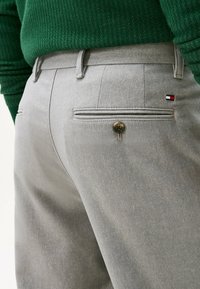 Un pantalon gris présente une texture lisse, des poches standard avec une fermeture à bouton, et une petite étiquette logo en rouge, blanc et bleu à l'arrière.