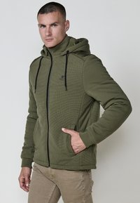 Alyvuogių spalvos zip-up džemperis, pagamintas iš tekstūruotos medžiagos, turintis gobtuvą, dvi šonines kišenes ir juodas raišteles. Logotipas ant krūtinės.