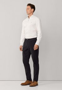 Witte lange mouwen overhemd met een geriefkraag met knopen, gecombineerd met een donkere broek en bruine suède schoenen, staand tegen een neutrale achtergrond.