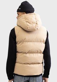 Chaleco acolchado beige con cuello alto y capucha, hecho de tela suave. Llevado sobre una camiseta negra de manga larga con textura, con un ajuste relajado.