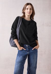 Maglietta henley a maniche lunghe nera con placca di bottoni, abbinata a jeans blu a gamba larga e una borsa a spalla grigia. Tessuto con texture e vestibilità casual.