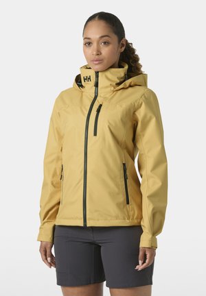 Vrouw draagt een waterdichte jas in mosterdgeel met capuchon en zwarte ritszakken, gecombineerd met donkergrijze korte broek, staand tegen een effen achtergrond.