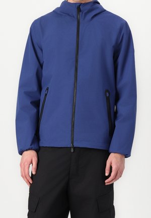 Mann trägt eine blaue Kapuzenjacke mit Frontreißverschluss und schwarzen Reißverschlusstaschen, kombiniert mit schwarzen Cargohosen.