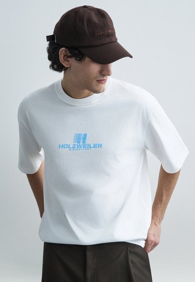 T-shirt blanc à manches courtes avec un logo graphique bleu "HOLZWEILER WINTERSPORT" sur la poitrine, associé à une casquette marron foncé.