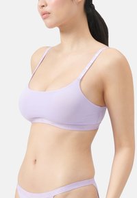 Frau in einem helllila Sport-BH und passender Unterwäsche, von den Schultern bis zu den Hüften zu sehen, mit zurückgebundenem Haar vor einem weißen Hintergrund.