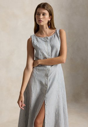 Donna che indossa un vestito senza maniche, abbottonato, lungo fino alla caviglia con strisce verticali blu e bianche, in piedi con un braccio incrociato sulla vita.