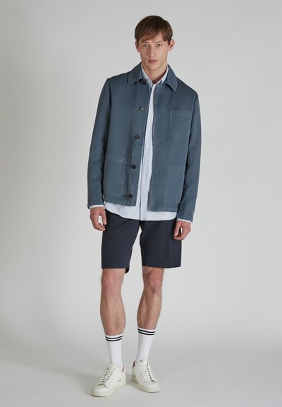 Giacca leggera blu con colletto, tre tasche frontali e chiusura con bottoni. Indossata sopra una camicia a righe bianche e pantaloni corti blu scuro. Sneaker bianche.