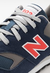 Modré semišové tenisky se šedými akcenty, bílým logem a červenými detaily. Mají šedé tkaničky a síťované boční panely, ukazující klasický design.