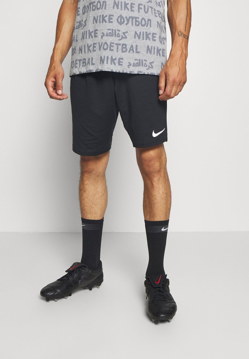 Shorts de sport noirs, T-shirt graphique gris avec du texte, chaussettes de football noires avec logo, et crampons noirs avec accents rouges.