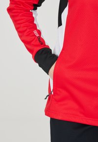 Giacca sportiva rossa con accenti neri e bianchi, tessuto testurizzato, maniche aderenti, dettaglio della tasca con zip sul lato.