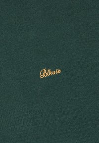 Σκούρο πράσινο πλεκτό ύφασμα με μικρό κεντημένο κίτρινο κείμενο "Blkvio" στο κέντρο.