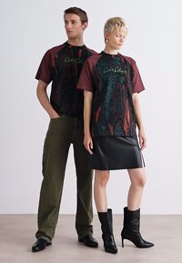 Twee modellen dragen grafische t-shirts met bordeauxrode korte mouwen, zwart en meerkleurige patronen. Vrouw in een zwarte leren rok en laarzen.