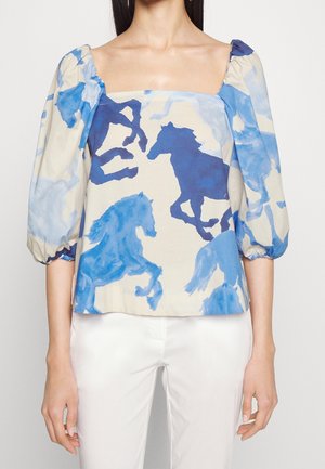 Femme portant un chemisier à col carré avec des manches bouffantes, avec des imprimés de chevaux bleus sur un fond crème, associé à un pantalon blanc.