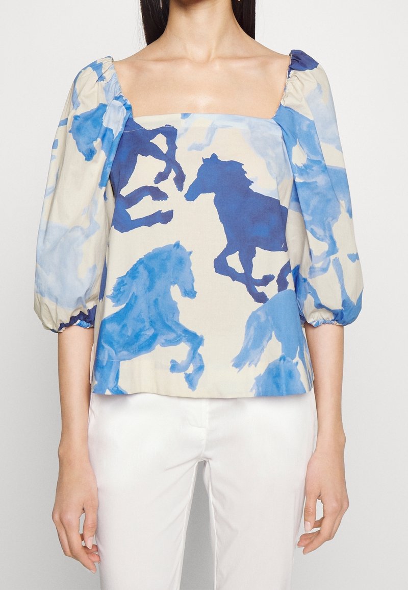 Femme portant un chemisier à col carré avec des manches bouffantes, avec des imprimés de chevaux bleus sur un fond crème, associé à un pantalon blanc.