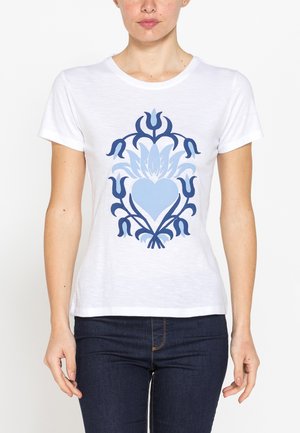 Donna che indossa una maglietta bianca a maniche corte con un disegno a cuore floreale blu, abbinata a jeans blu scuro, in piedi contro uno sfondo semplice.