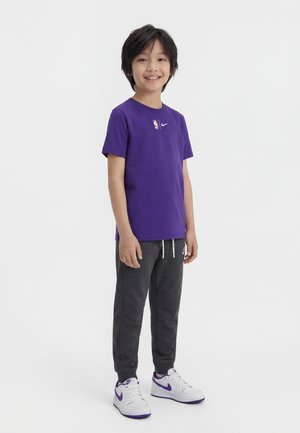 Dijete stoji i smiješi se, nosi ljubičastu NBA majicu, tamnosive trenirke i bijele tenisice s ljubičastim detaljima.