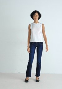 Hvit utenermet topp med dekorative detaljer, parret med mørkeblå rette jeans og svarte flate sko. Enkel, uformell design.