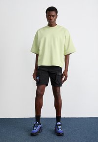 Light green oversized sweatshirt, black cargo shorts with patches, and blue athletic shoes. Smooth fabric, relaxed fit, and short sleeves.