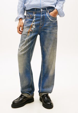 Persona que lleva puestos unos vaqueros azules desgastados de corte holgado con una cadena plateada decorativa y zapatos de cuero negros, de pie con una mano en el bolsillo.