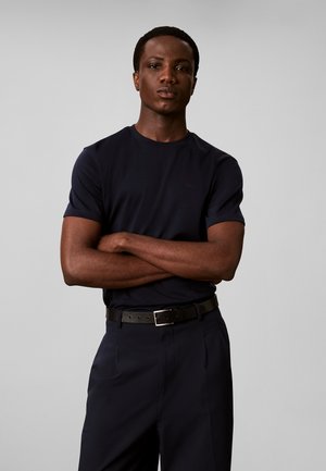 Hombre con los brazos cruzados, vistiendo una camiseta negra y pantalones negros, de pie frente a un fondo liso de color gris claro.