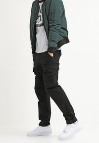 Groene bomberjack, grijze T-shirt met logo, zwarte cargobroek en witte sneakers. Effen kleuren, slim-fit, gestructureerde materialen en meerdere zakken.