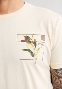 Camiseta color crema hecha de tela suave, con un estampado botánico de hojas verdes y un diseño de texto marrón en el frente.