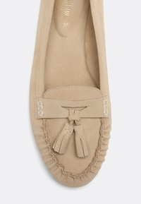 Mocassin en daim beige avec un bout arrondi, des détails cousus, un nœud à pompons sur le dessus, et le nom de la marque visible à l’intérieur du talon.